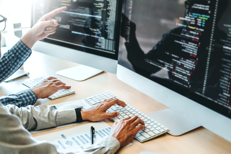 two colleagues looking over code on large desktop computer screens